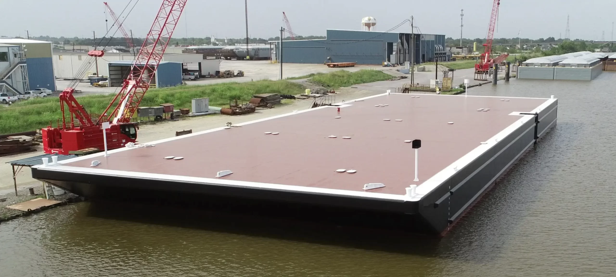 Large flat-deck barge floating at an industrial shipyard with cranes and equipment nearby