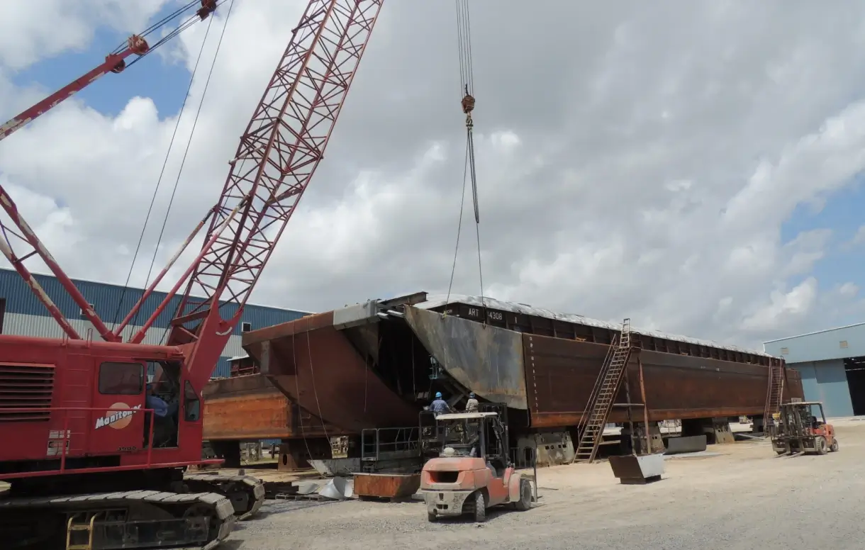 Large steel barge section being lifted by a crane at an industrial shipyard site.