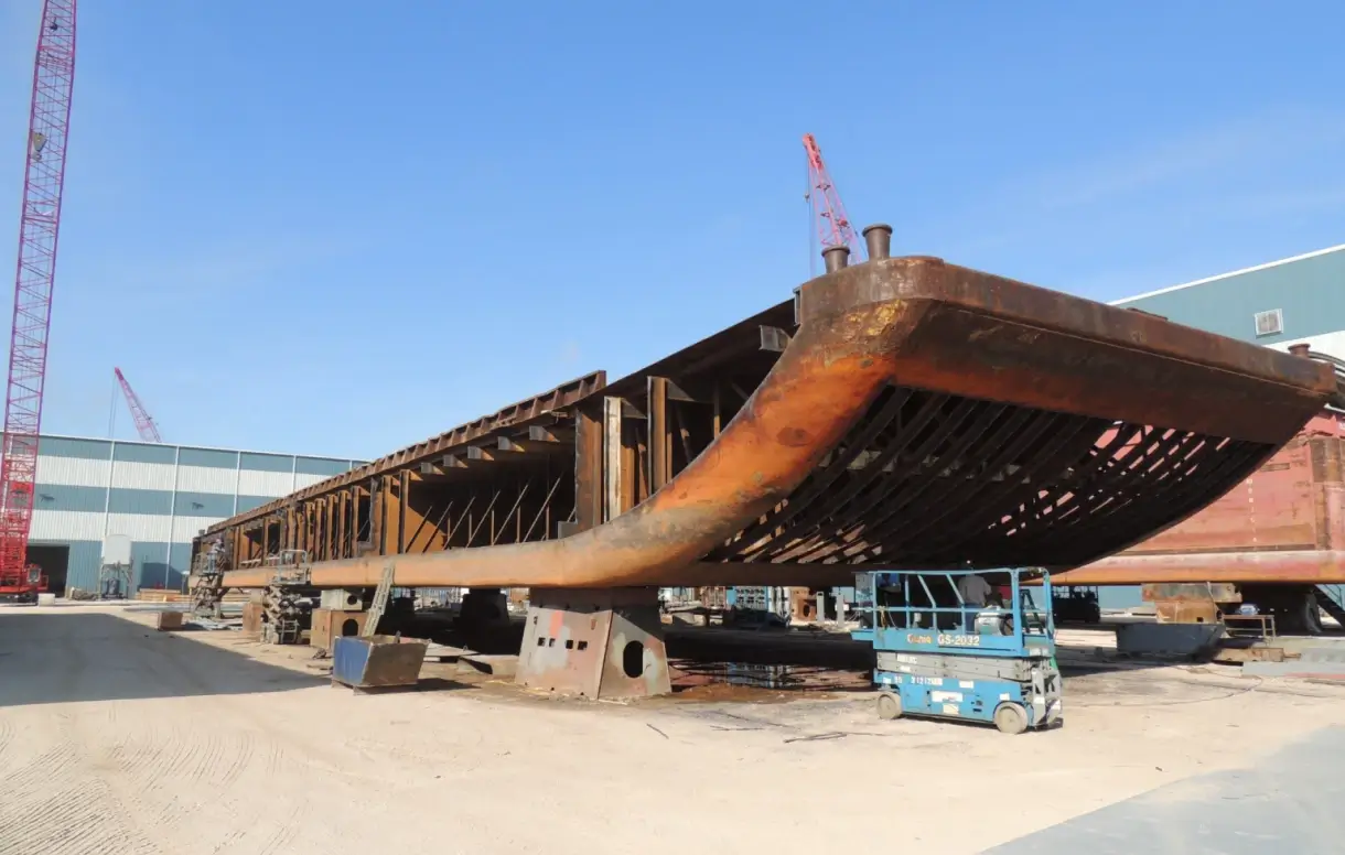 Large steel barge under construction on supports at a shipyard with scaffolding and equipment underneath.