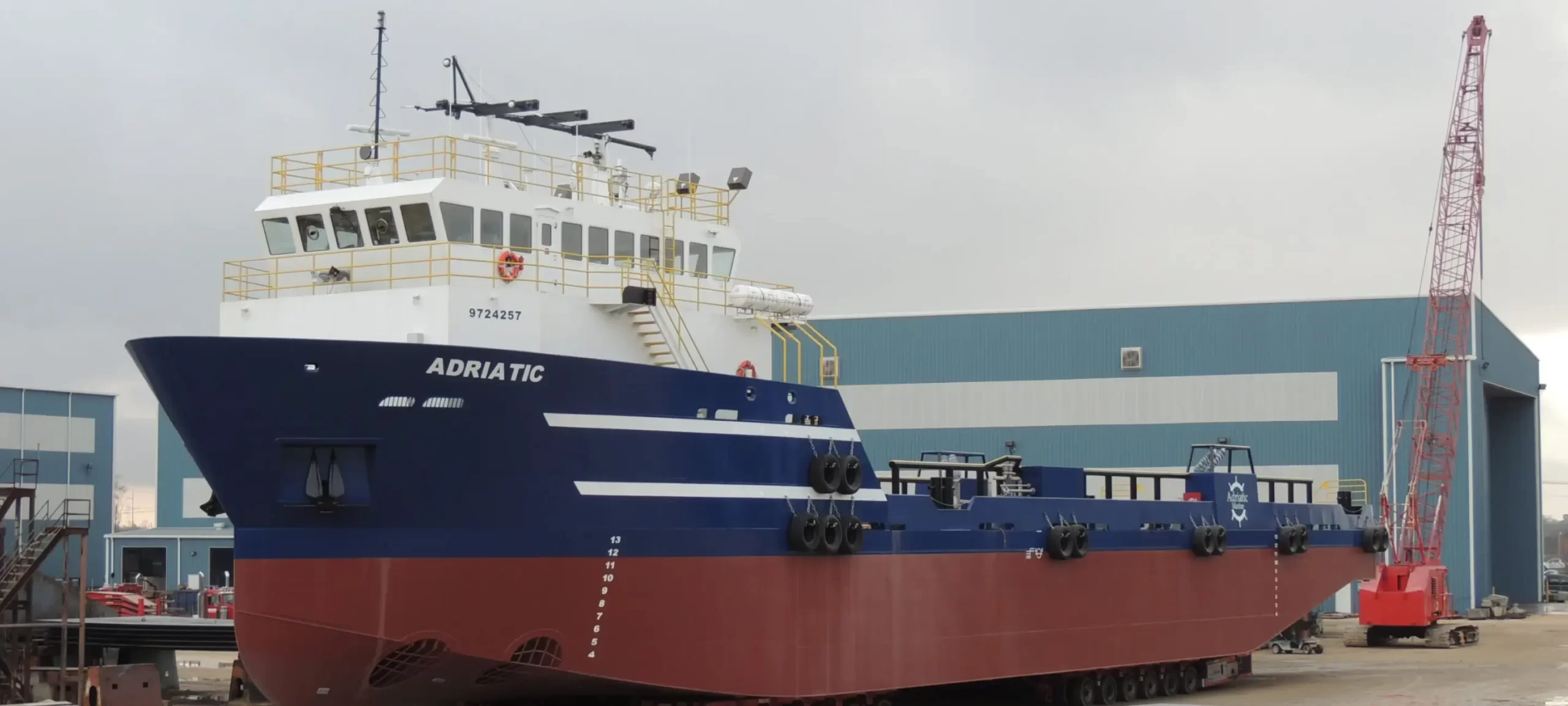 White, blue and red offshore support vessel named Adriatic docked beside a shipyard building.