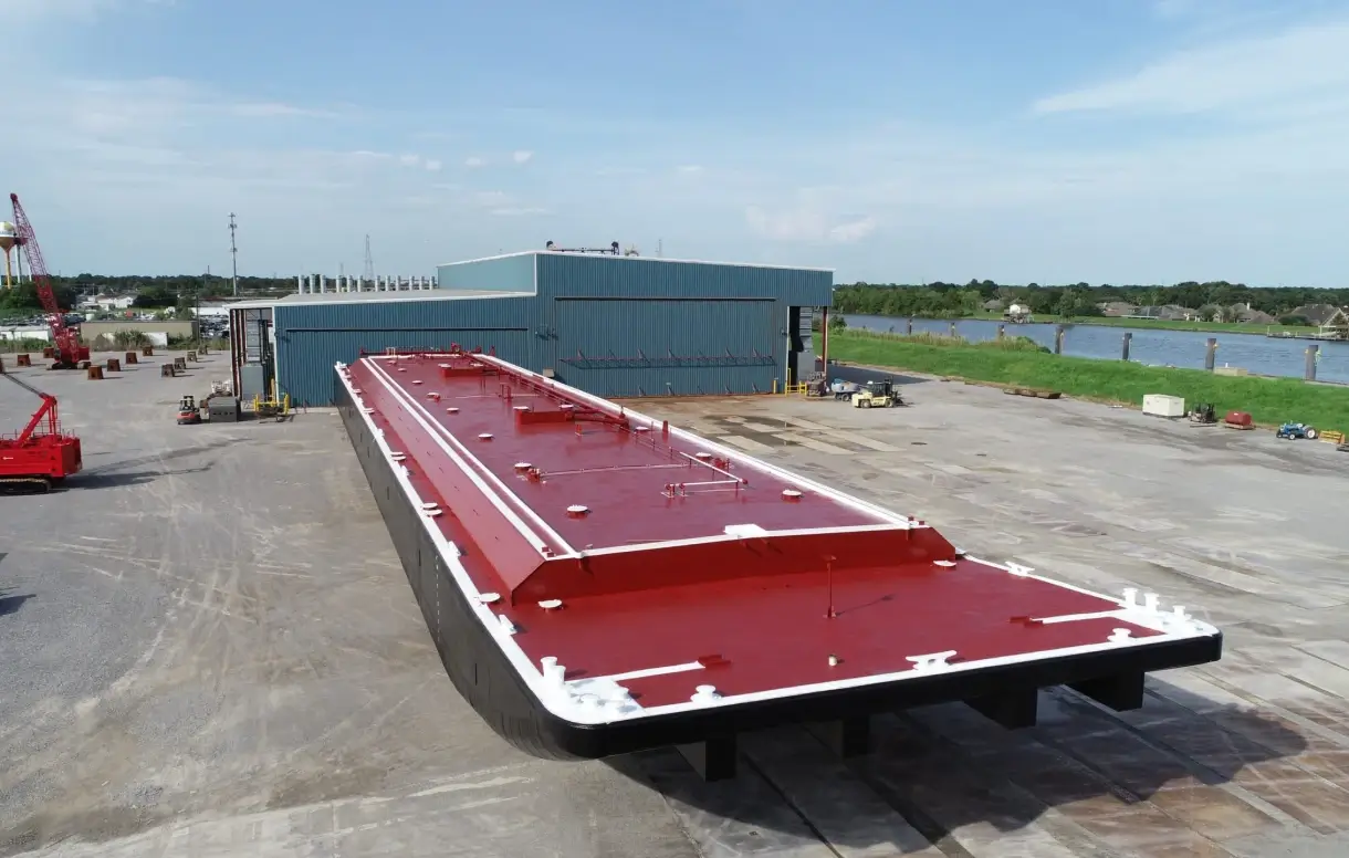 Newly built flat-deck barge on land at a shipyard with red-painted deck and white edging.
